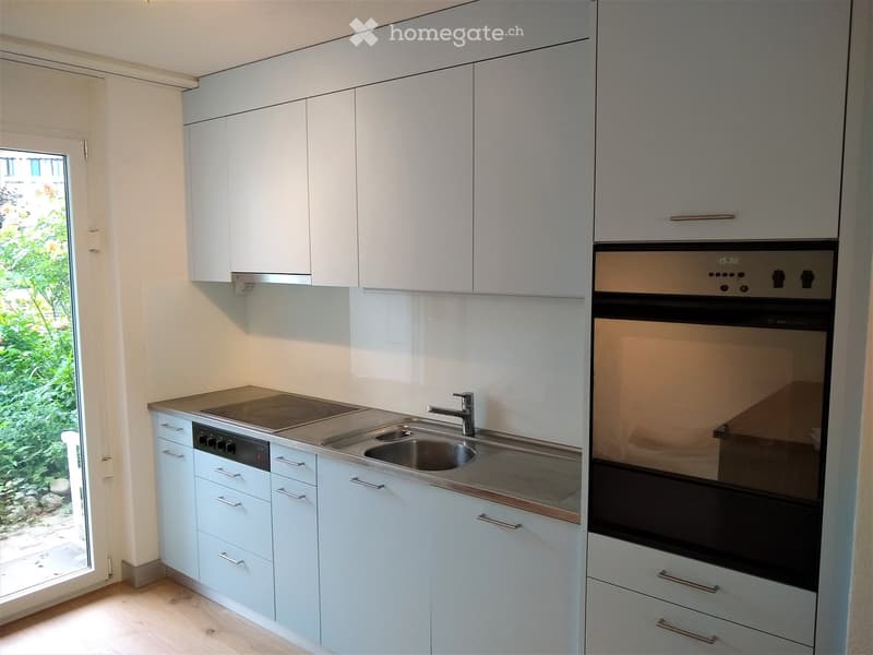 Modern kitchen with white cabinets, stainless steel sink, oven, and stove. Features a large window with a view of greenery outside.