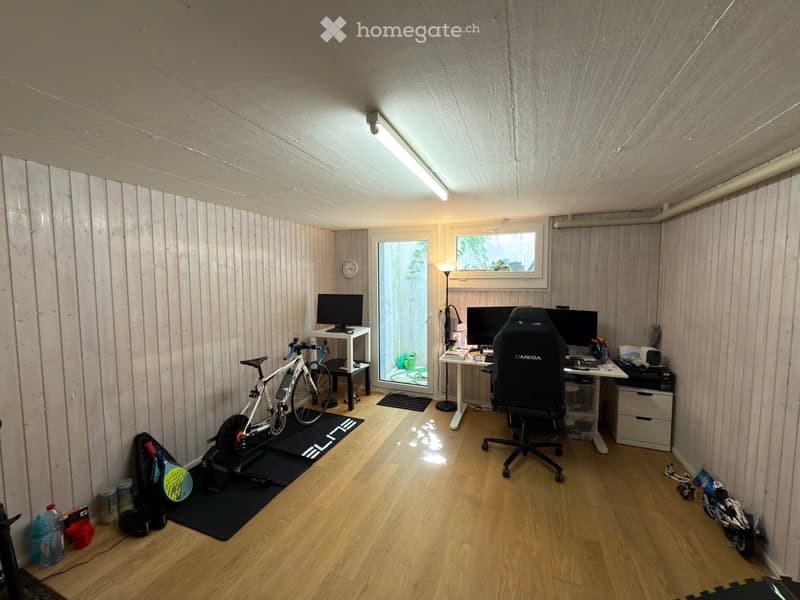 A well-lit room with wooden walls and flooring, featuring a desk with two computer monitors, a gaming chair, a bicycle, and a small table with a clock.