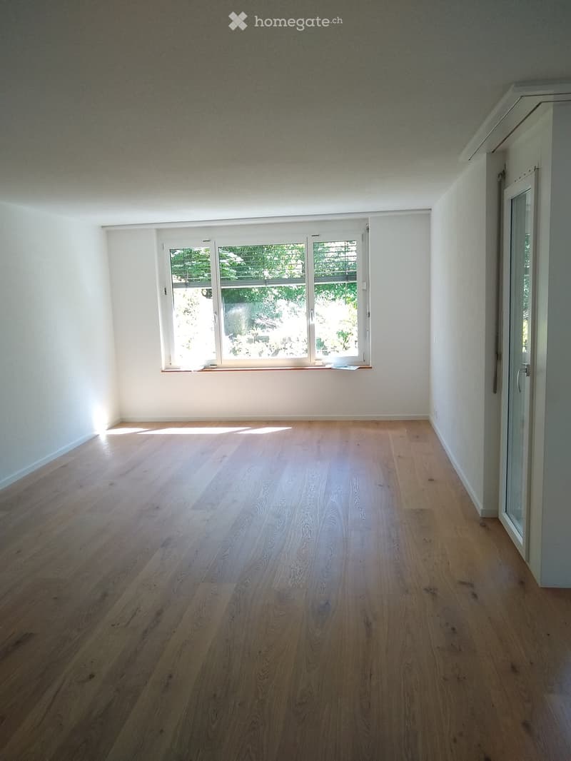 Empty room with light wooden flooring and white walls, featuring a large window with blinds and a door leading to another space.
