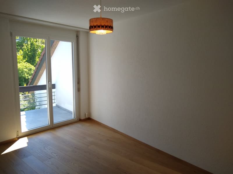 Empty room with wooden flooring, white walls, large sliding glass door leading to a balcony, and a hanging lamp.