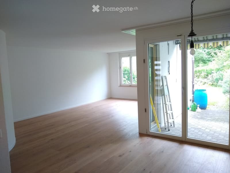 Empty room with wooden flooring, white walls, large glass sliding doors leading to an outdoor area, and a window on the left side.