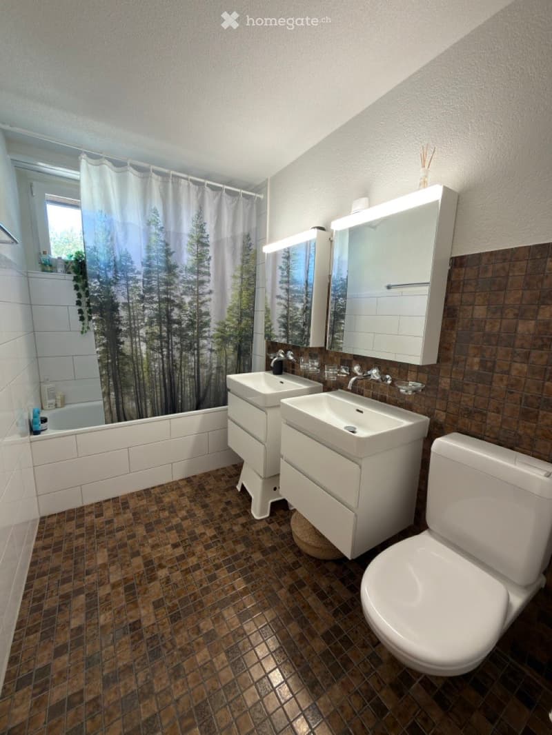 Modern bathroom with a double sink vanity, toilet, bathtub, and a large mirror with built-in storage. The floor features brown tiles, and the walls have a mix of white and brown tiles. A forest-themed shower curtain is visible in the background.