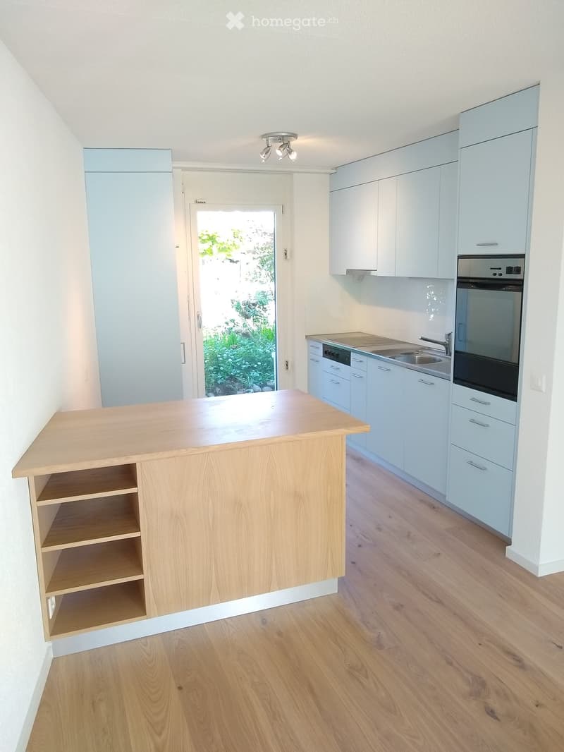 Modern kitchen with light blue cabinets, wooden island, sink, and oven. The space features wooden flooring and a view of greenery through a glass door.