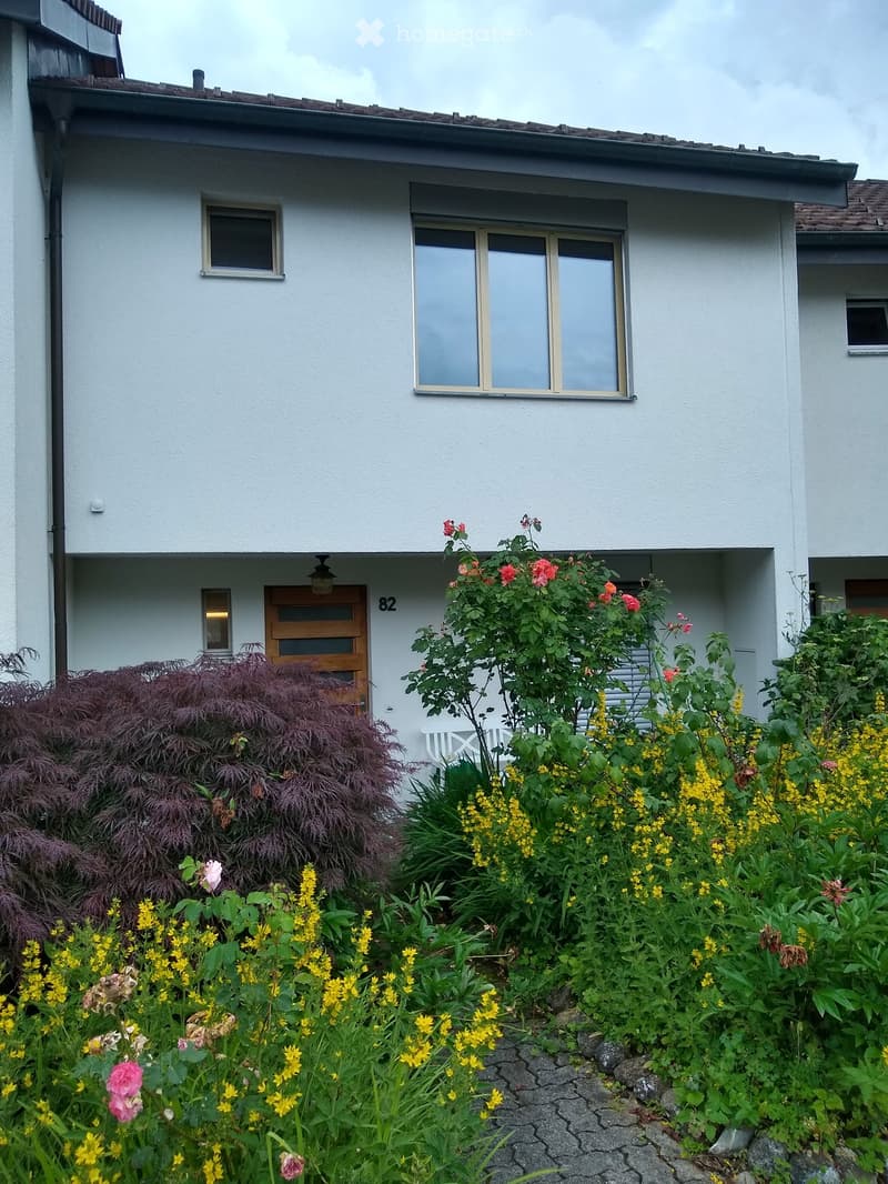 Front garden with colorful flowers, shrubs, and a small white bench near the entrance of a two-story house.