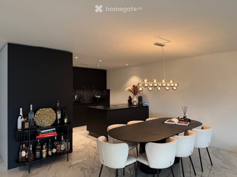 Modern kitchen and dining area with a round dining table, white chairs, a dark cabinet, and a shelf with bottles and decorative items.