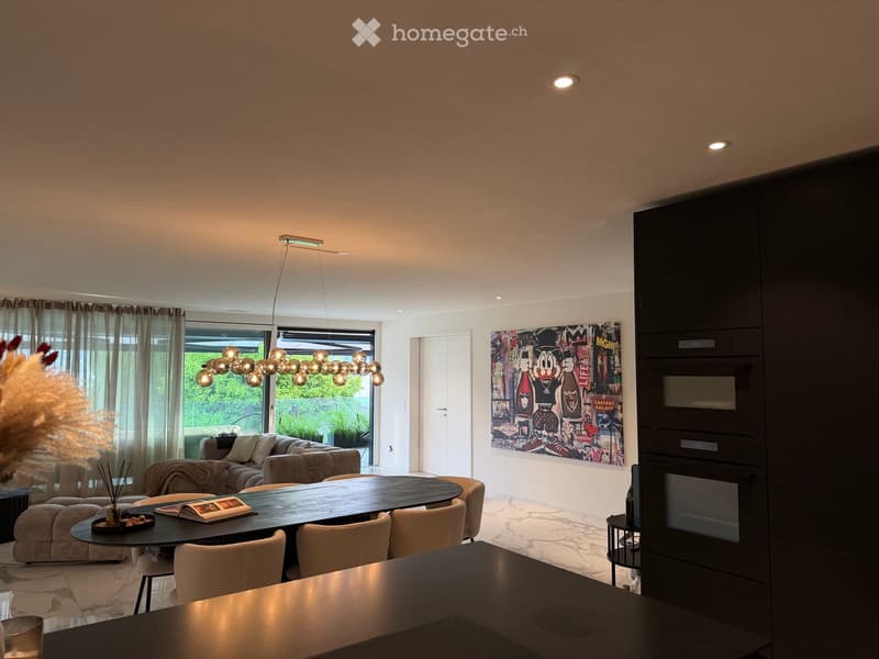 Modern living room with a round dining table, chairs, sofa, decorative lamp, and artwork on the wall. Large windows with curtains provide natural light.