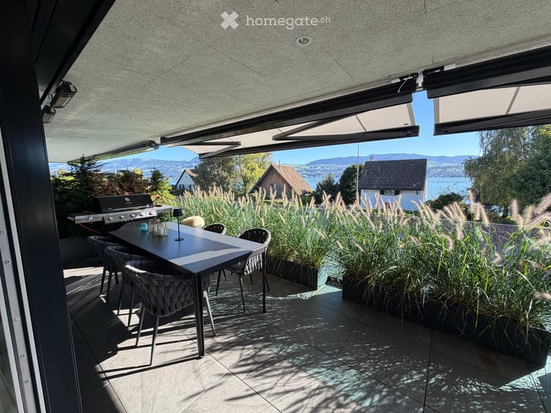 A modern covered outdoor terrace with a dining table, chairs, and a grill, overlooking a scenic lake and mountains.