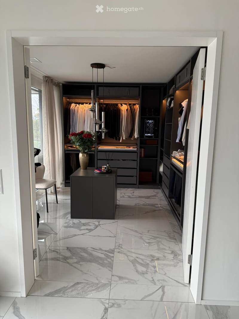 Elegant hallway with marble flooring, modern built-in wardrobes, a small black console table with a vase of red flowers, and a sleek hanging light fixture.