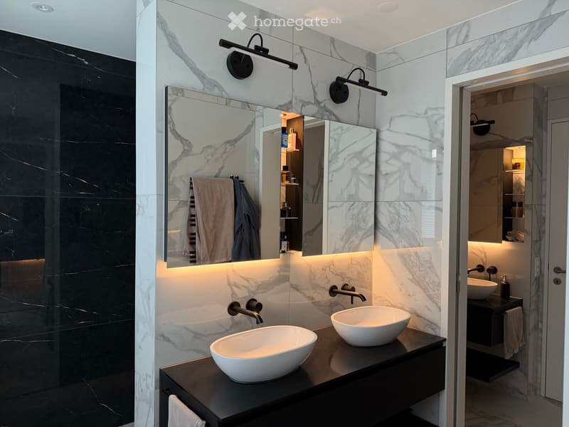 Modern bathroom with marble walls, two white sinks, sleek black fixtures, illuminated mirror, and built-in storage.