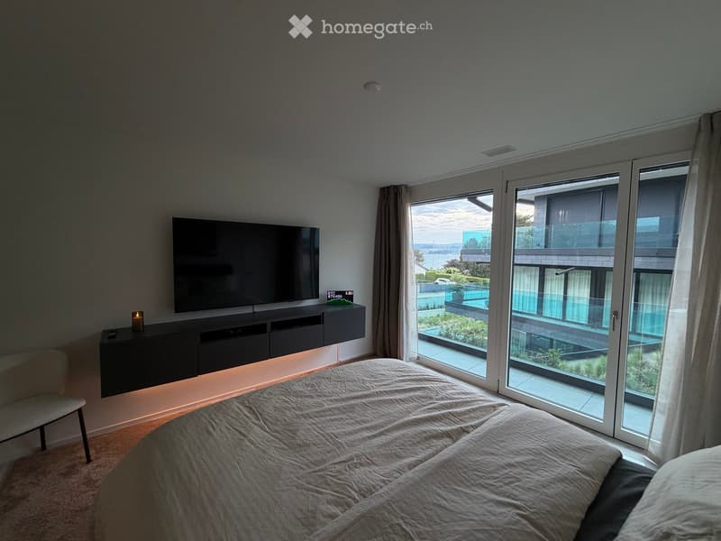 Modern bedroom with a large bed, a TV mounted on the wall, a sleek black TV stand, a small chair, and large windows offering a scenic view of the water and surrounding area.