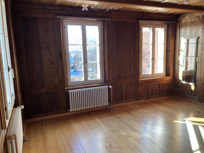Empty room with wooden walls and floor, large windows, radiators, and exposed wooden beams on the ceiling.
