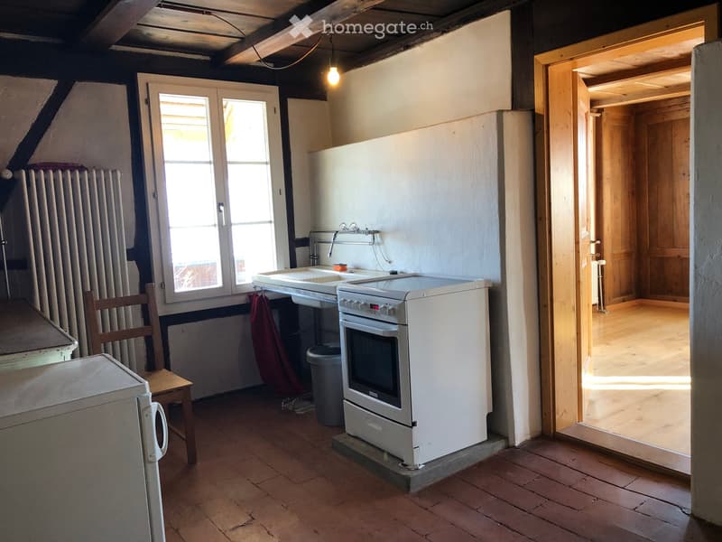 Small kitchen with exposed wooden beams, brick floor, sink, faucet, oven, refrigerator, and a wooden chair near the window.