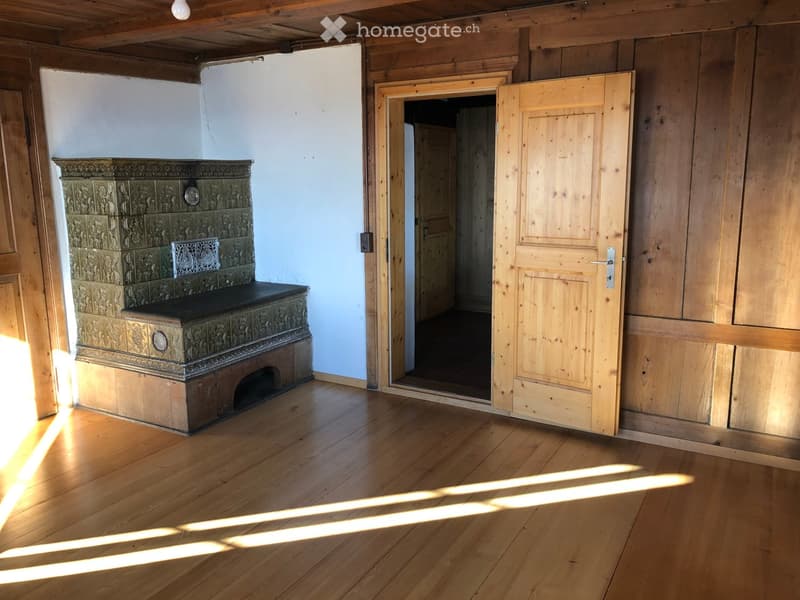 Empty room with wooden walls, wooden floor, and a large ornate fireplace. Sunlight streams in from a window, casting shadows on the floor.