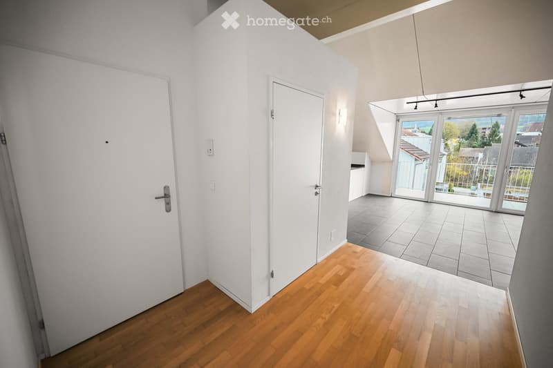 Bright hallway with wooden flooring, white walls, and two doors leading to other rooms. A large window with a view of the outside area is visible at the end of the hallway.
