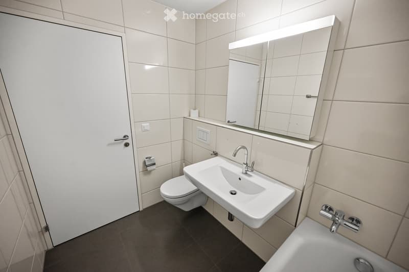 Modern bathroom with white tiles, a sink with a chrome faucet, a toilet, a bathtub, and a large mirrored cabinet with built-in lighting.