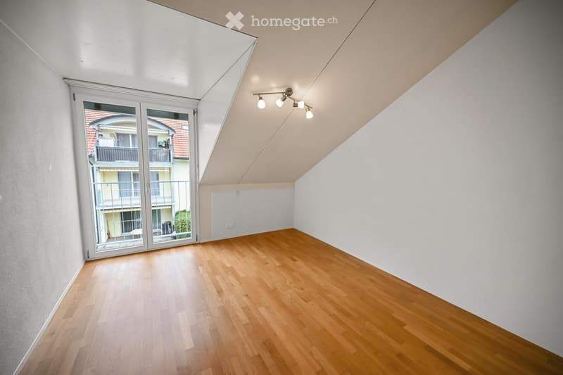 Empty room with wooden flooring, white walls, and a large window with a view of a neighboring building and balcony.