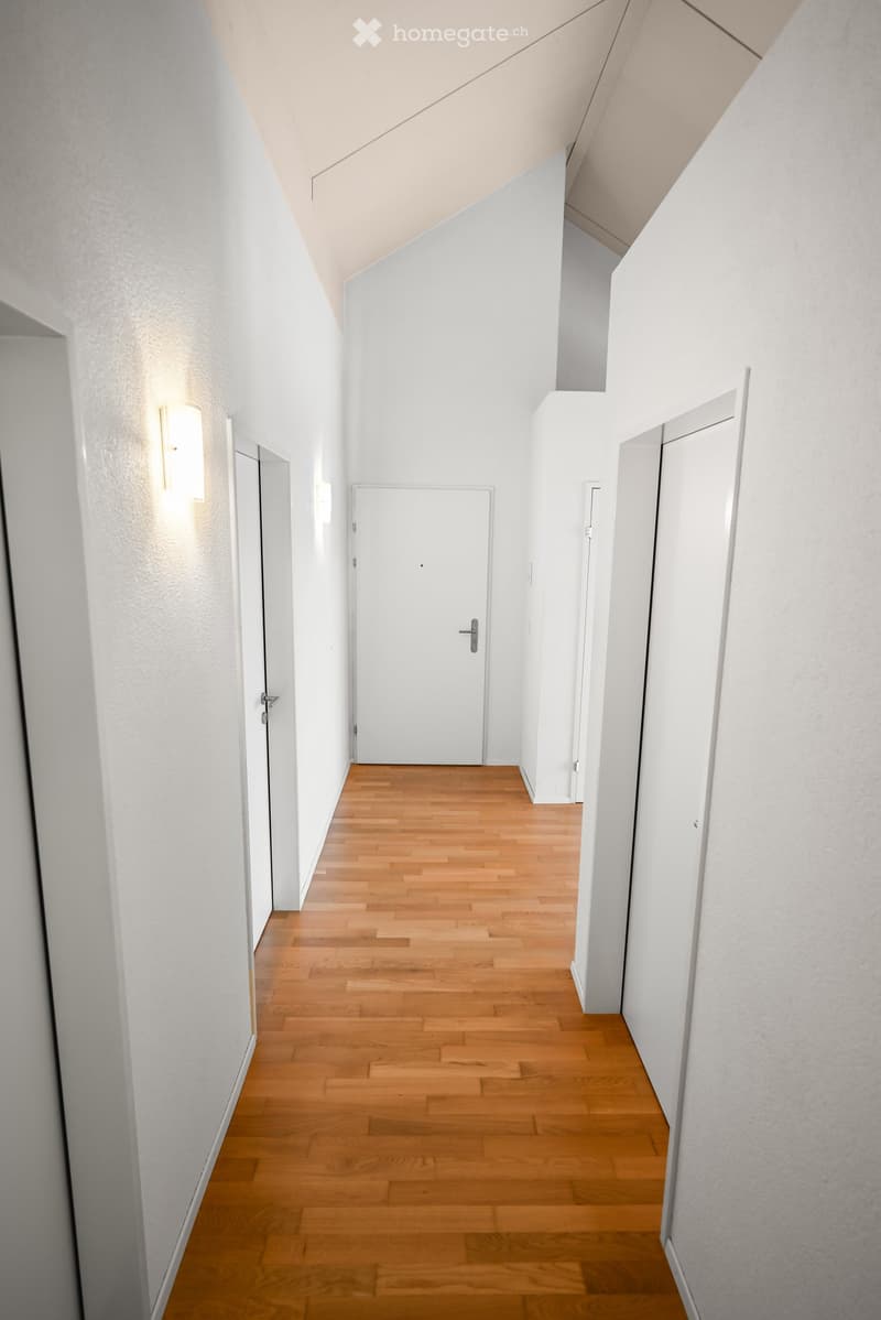 A clean, modern hallway with light wooden flooring, white walls, and several closed doors. The ceiling has a sloped design, and there are wall-mounted light fixtures on either side.