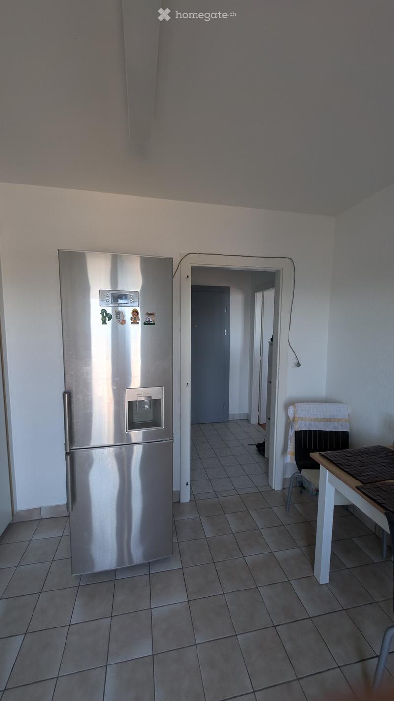 A small kitchen with a stainless steel refrigerator, a small table with a chair, and a tiled floor. The room has white walls and a doorway leading to another room.