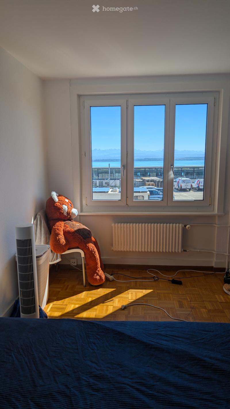 Bright bedroom with wooden flooring, large window offering a sea view, bed with blue bedding, fan, radiator, chair, and a large stuffed animal.