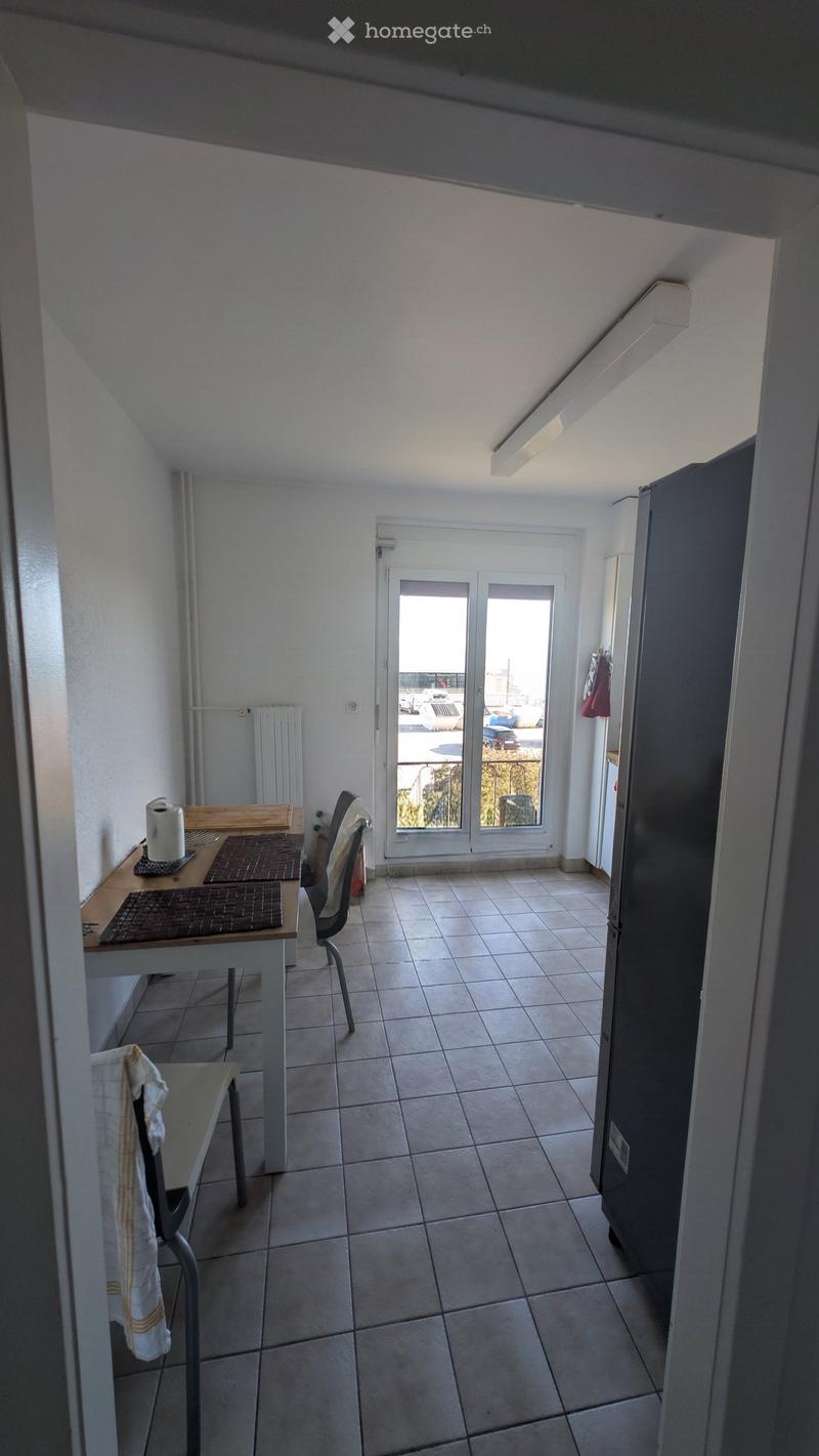 A small, well-lit room with tiled flooring, a wooden table with chairs, a refrigerator, and a sliding glass door leading outside.