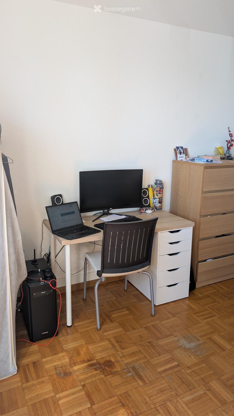 A modern, well-lit study or home office with a wooden parquet floor, a white desk with a laptop, screen, keyboard, mouse, and speakers, a black office chair, a white drawer unit, and a black tower computer on the floor.