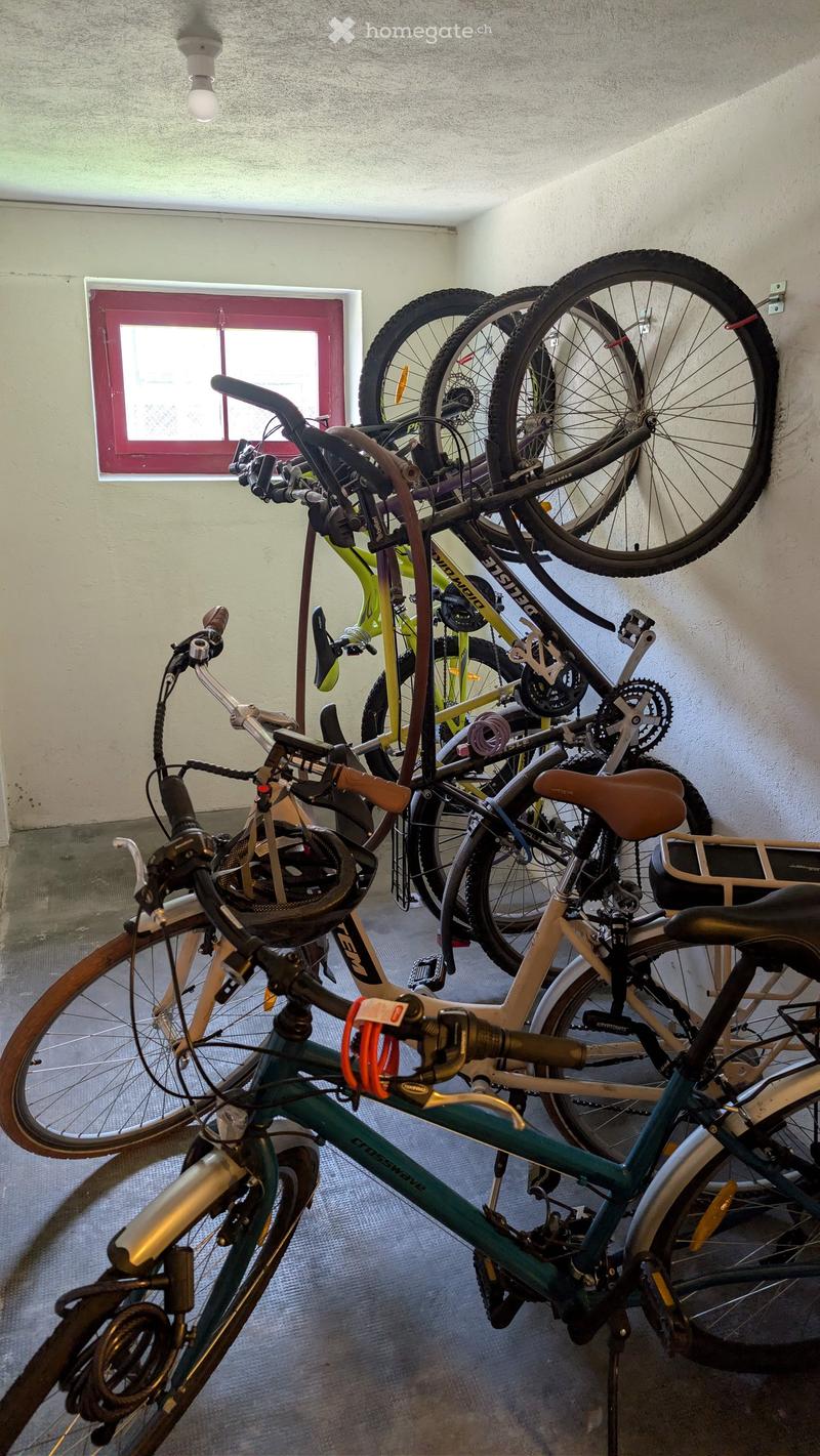 A small, well-lit room with several bicycles stored inside, including a mix of road and mountain bikes, with a window and a door visible.