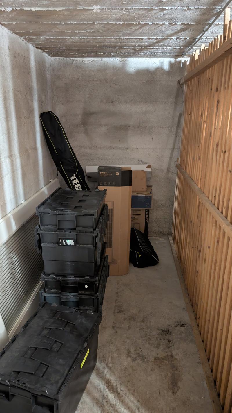 A narrow, unfinished hallway with concrete walls and a wooden railing on one side. Various storage items, including black plastic bins, cardboard boxes, and a folded item, are stacked along the left wall.