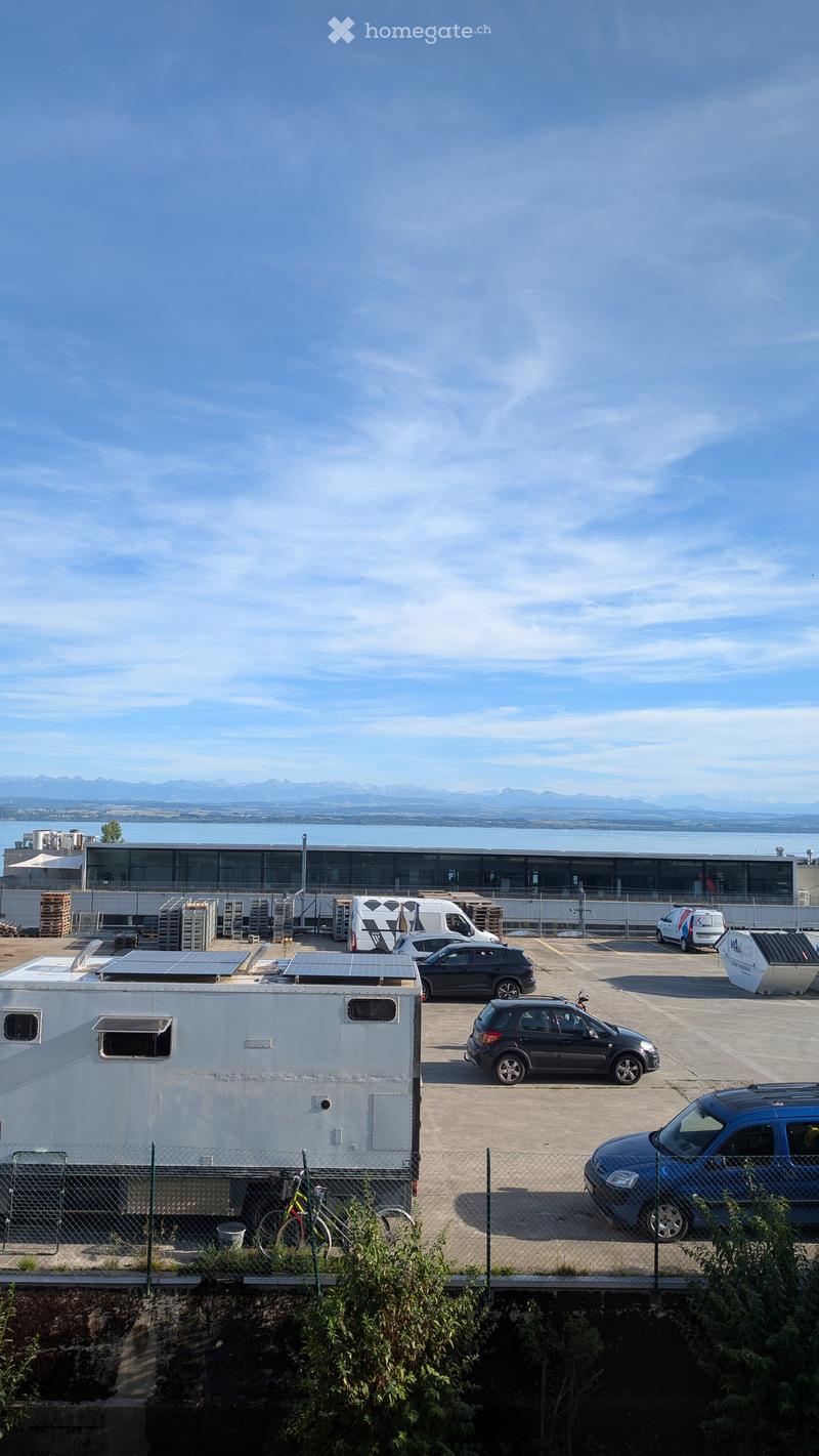 Parking lot with several vehicles, trailers, and a fenced area with a bicycle and solar panels, overlooking a body of water and distant mountains.