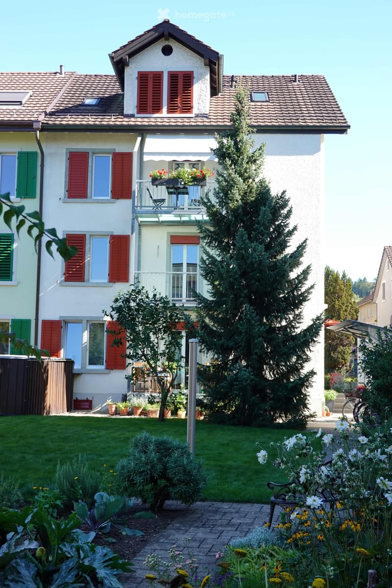 A well-maintained garden with a variety of plants, flowers, and a small seating area near the house. The house features multiple windows with shutters, a balcony, and a neatly trimmed lawn.