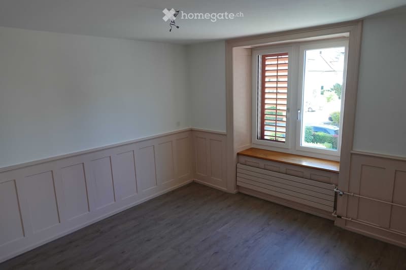 Empty room with light blue walls, wooden flooring, and a large window with blinds. The room features built-in paneling and a radiator beneath the window.