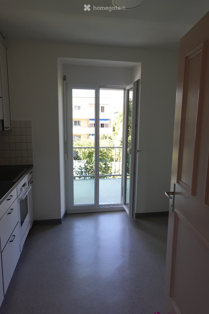 Empty hallway with white cabinets on one side, a sliding glass door leading to a balcony, and a view of greenery outside.