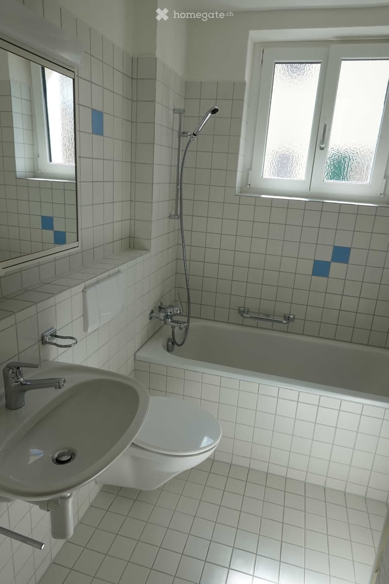 Bathroom with white tiled walls and floor, featuring a sink, toilet, bathtub, and showerhead. Windows provide natural light.