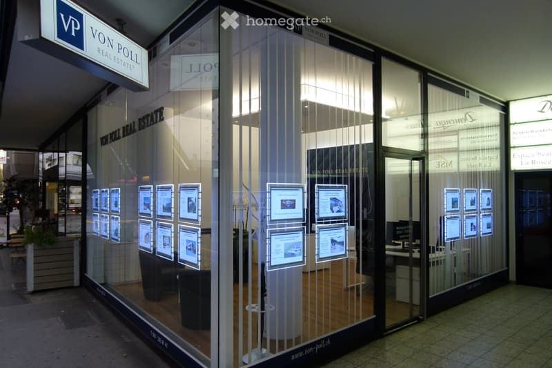 Exterior view of a real estate office with glass windows displaying property listings, signage for "Von Poll Real Estate", and interior furniture visible through the glass.