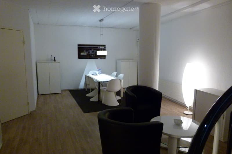 Modern, minimalist room with a white dining table and chairs, a black armchair, a tall floor lamp, and a small cabinet with bottles on top.