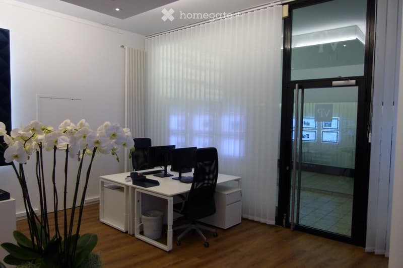 Modern office space with a white desk, black office chair, two computer screens, and a vase of white orchids. The room features large windows with white curtains and a glass door leading to another area.