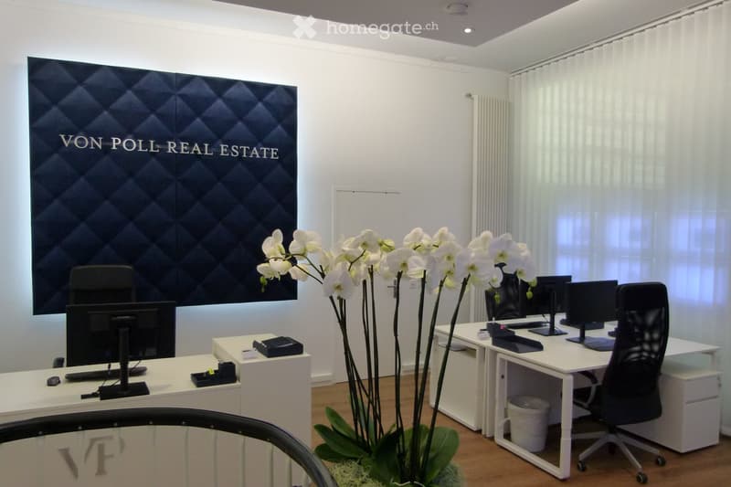 Modern office space with white desks, black chairs, multiple computer screens, and a large flower arrangement in the center.