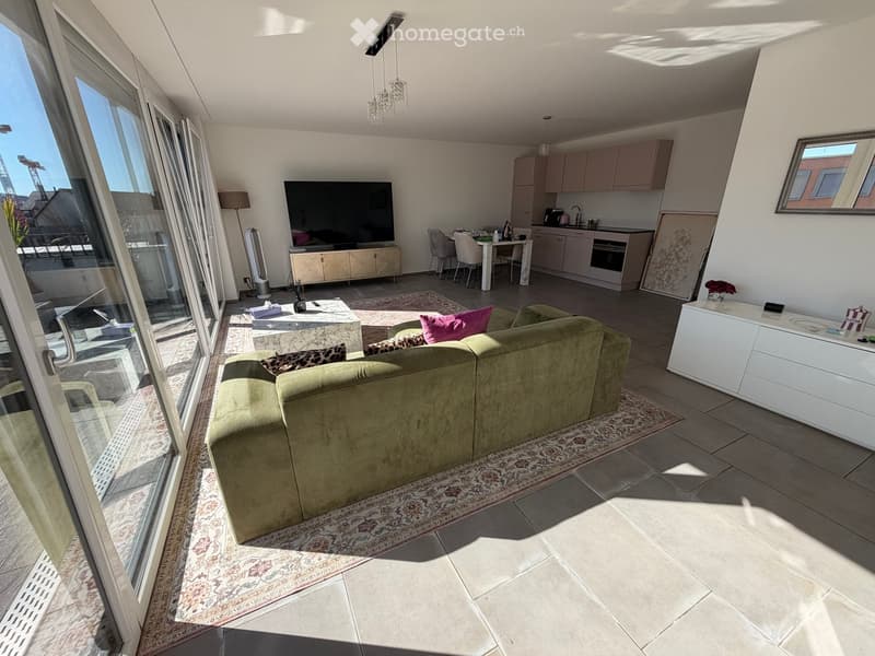 Bright living room with a green sofa, coffee table, TV, and a small dining area with chairs and a table. The kitchen area features light-colored cabinets, an oven, and a sink with a faucet. Large windows offer a view of the outside.