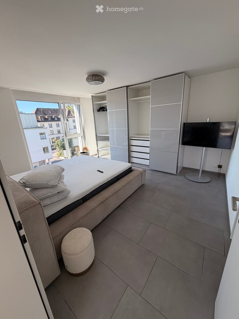 Modern bedroom with a large bed, a round stool, a wardrobe, and a TV on a stand. Large windows offer a view of the street.