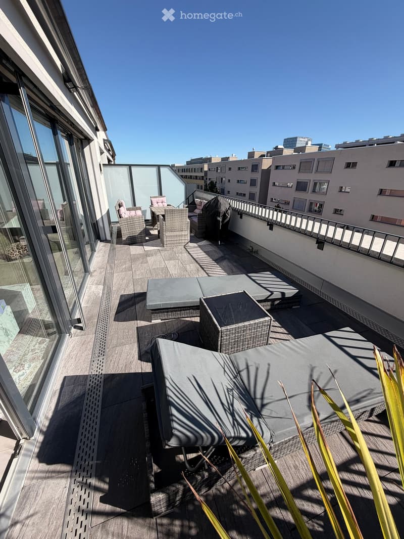 Balcony with wicker chairs, a table, and a grey sofa, overlooking a cityscape with modern buildings.