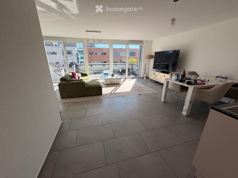 Modern living room with a green sectional sofa, coffee table, dining table with chairs, and a large flat-screen TV. The room features large windows with a view of a balcony area.