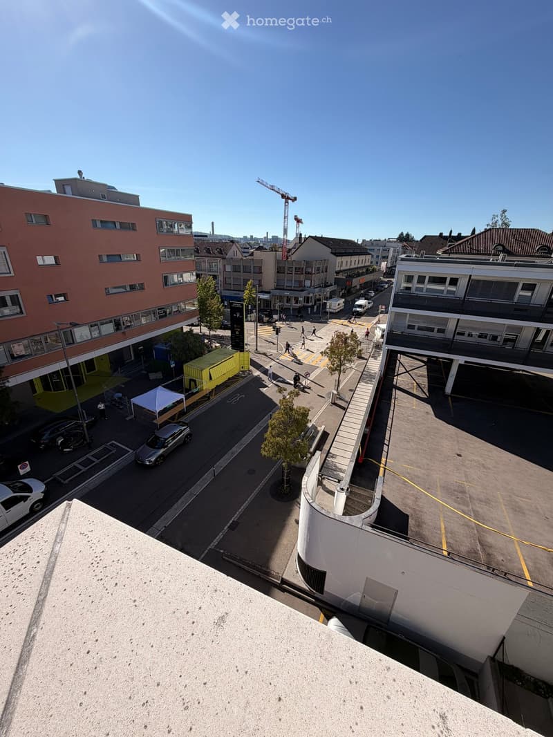 Aerial view of an urban area with multi-story buildings, a parking lot, a few cars, pedestrians, and a small green tent setup.