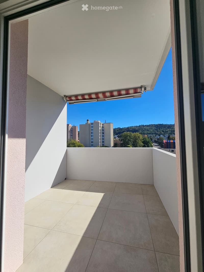 Balcony with tiled floor, white walls, and a retractable awning, offering a view of buildings and greenery.