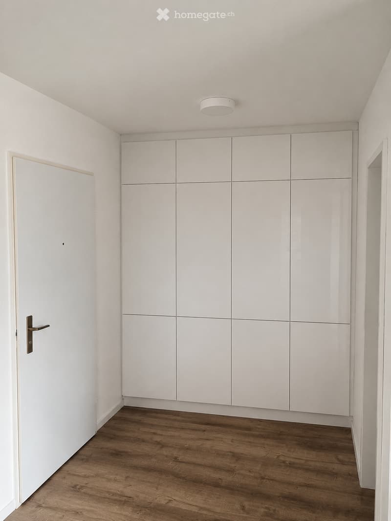 An empty room with white walls, wooden flooring, and built-in white cabinetry. A single door is visible on the left side, and a recessed ceiling light is mounted on the ceiling.