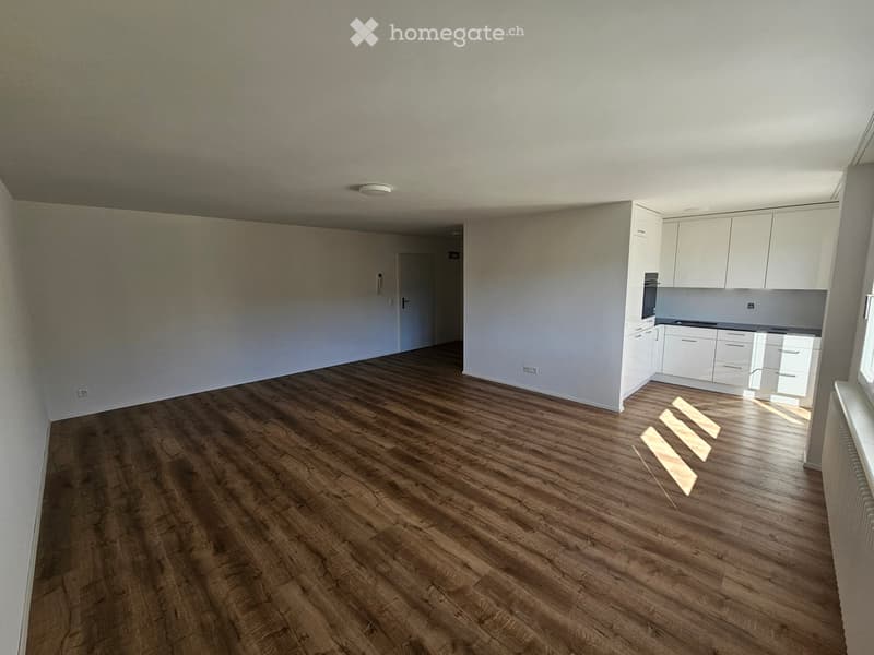 Empty room with wooden flooring, white walls, and a small kitchen area with white cabinets and appliances.