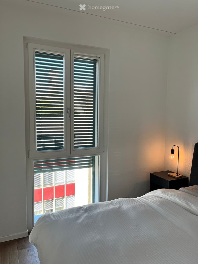 Modern bedroom with a neatly made bed, nightstand, and a wall-mounted lamp. Large windows with blinds offer natural light and a view of the outside.