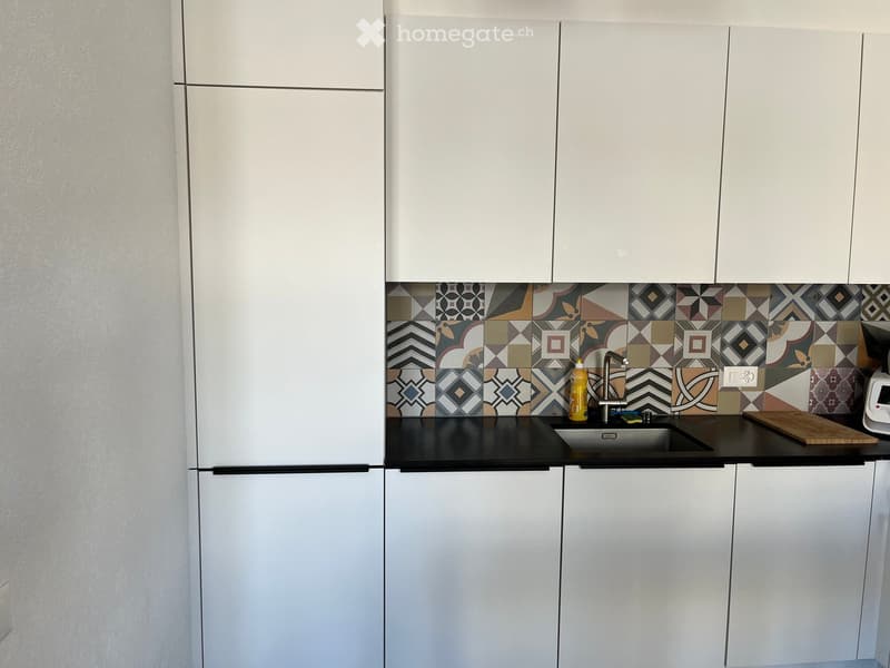 Modern kitchen with white cabinets, black countertop, and colorful geometric tile backsplash. Features a sink, faucet, bottle, cutting board, and timer.