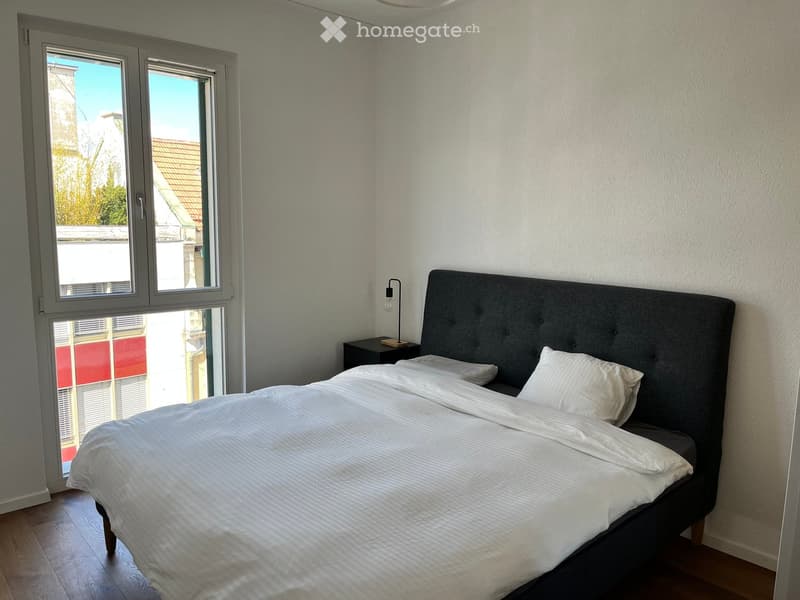 Modern bedroom with a grey upholstered headboard, white bedding, a nightstand with a lamp, and large windows offering a view of the street.