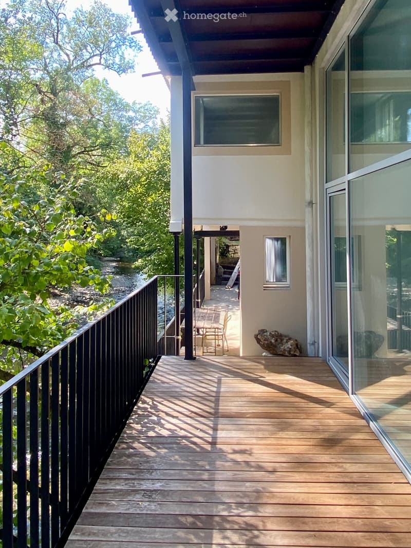 Wooden balcony with black railing, overlooking a lush green area with trees and a small stream. A table and chairs are placed on the balcony.