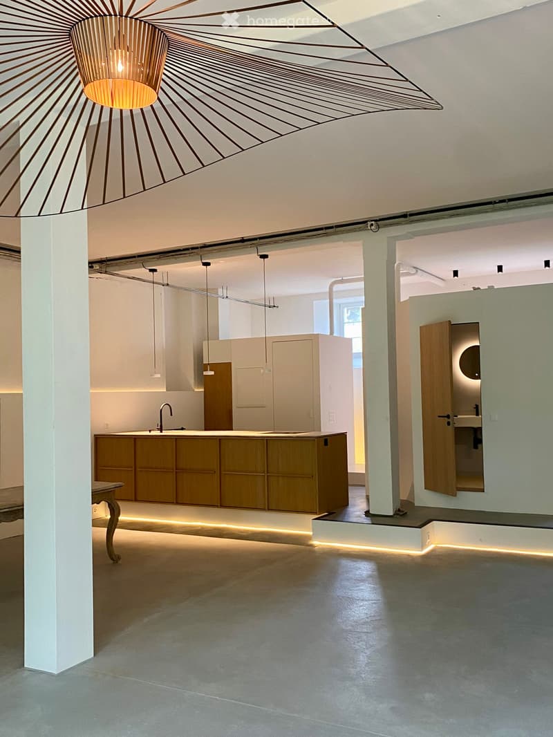 Open-plan space with a modern kitchen area, a sink, and a table, illuminated by a geometric pendant light and recessed lighting.