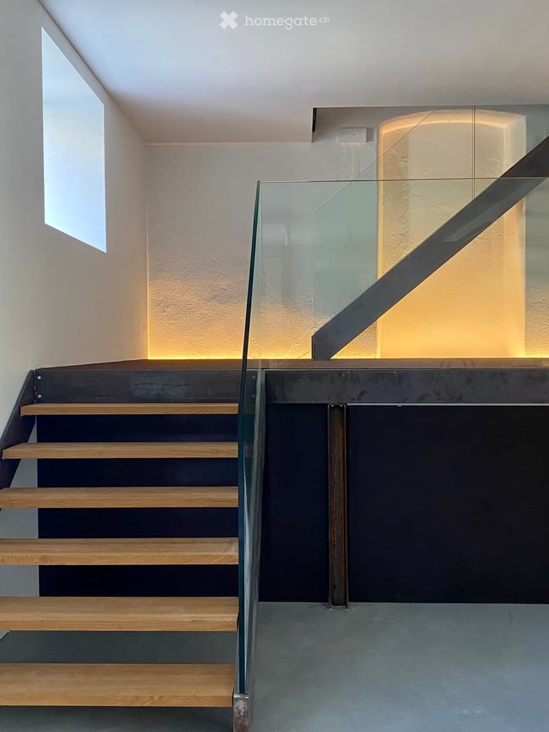 Modern staircase with wooden steps and a glass railing, illuminated by warm lighting on the walls.